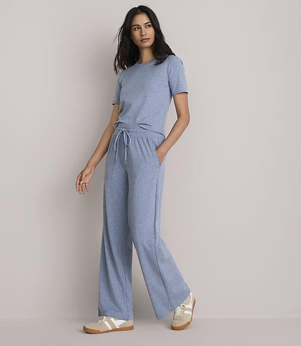 A woman in a blue t-shirt and wide-leg pants stands against a plain, light gray background.