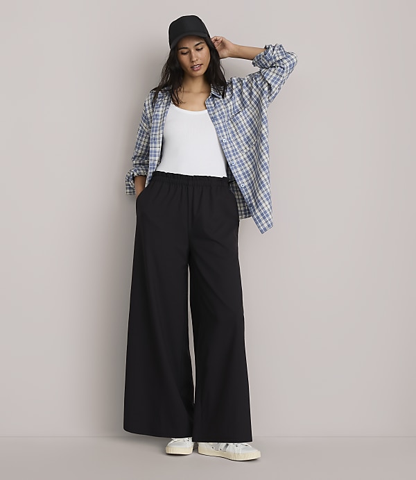 A woman in a white tank top, black wide-leg pants, and an open plaid shirt stands against a plain gray background.