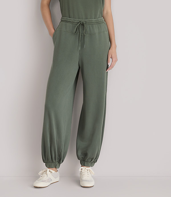 A person wearing olive green sweatpants and sneakers stands against a plain white background.