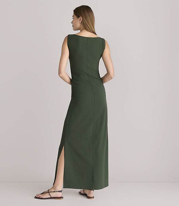 A woman in a long, dark green dress with a thigh-high slit stands on a white floor against a plain white background.