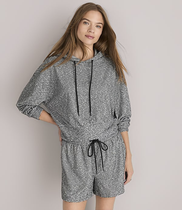 A woman in a grey patterned hoodie and shorts sits against a plain grey background.