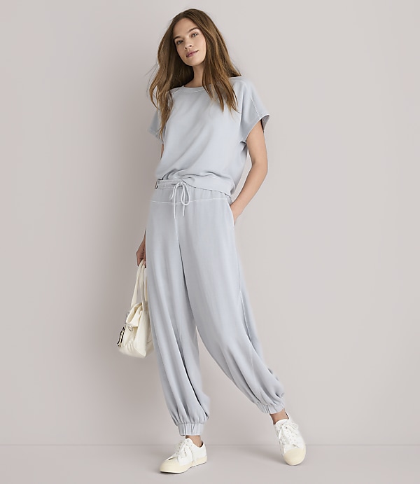 A woman in a light blue loungewear set and white sneakers stands against a plain white background, holding a cream-colored tote bag.