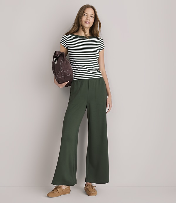 A woman in a striped shirt and wide-leg green pants stands against a plain white background, holding a brown bag.