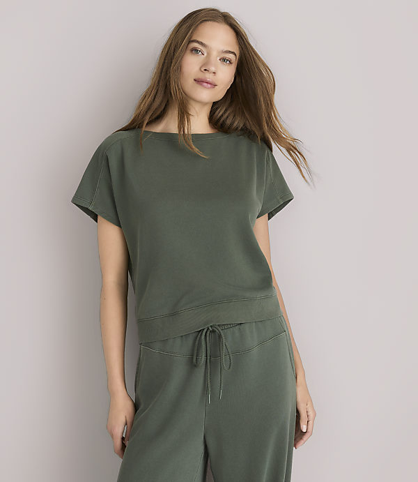 A young woman in a green top and pants stands against a plain gray background.