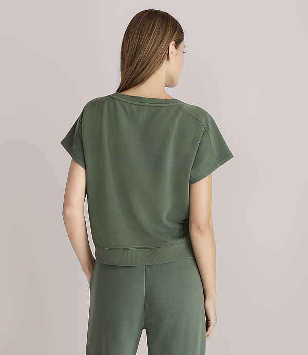 A woman in a green t-shirt and pants stands against a plain gray background.
