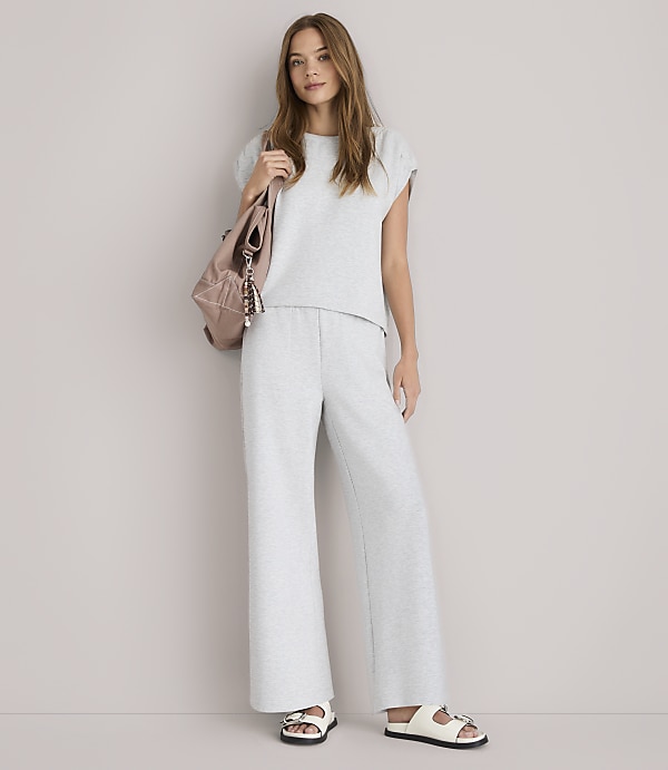 A young woman in a grey loungewear set and white sandals stands against a plain white background, with a pink backpack slung over her shoulder.