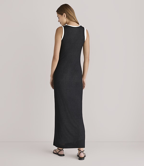 A woman in a black maxi dress with a white trim stands facing away from the camera against a plain, light-colored background.