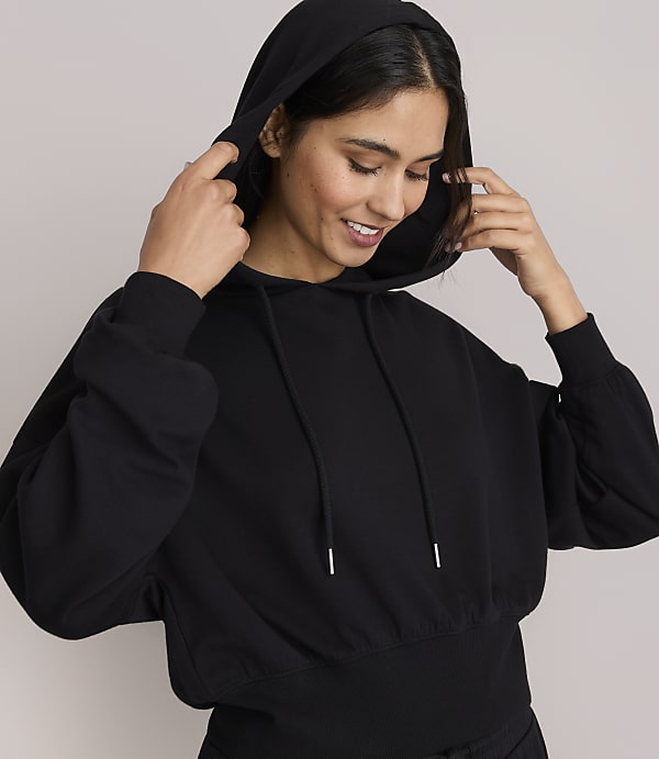 A smiling woman in a black hoodie adjusts the hood against a plain gray background.