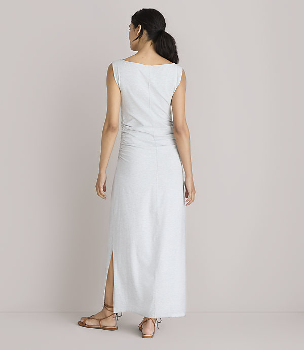 A woman in a light gray maxi dress with ruched detailing and a side slit stands with her back to the camera against a plain white background.