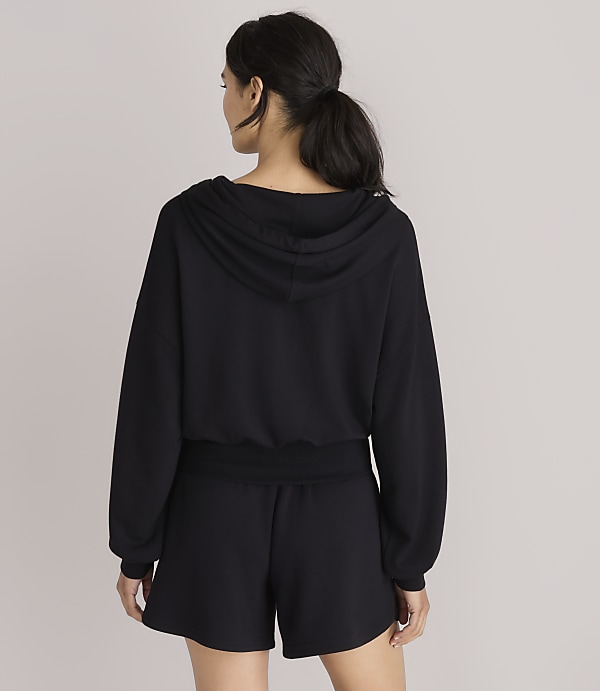 A woman in a black hoodie and shorts stands against a plain white background.