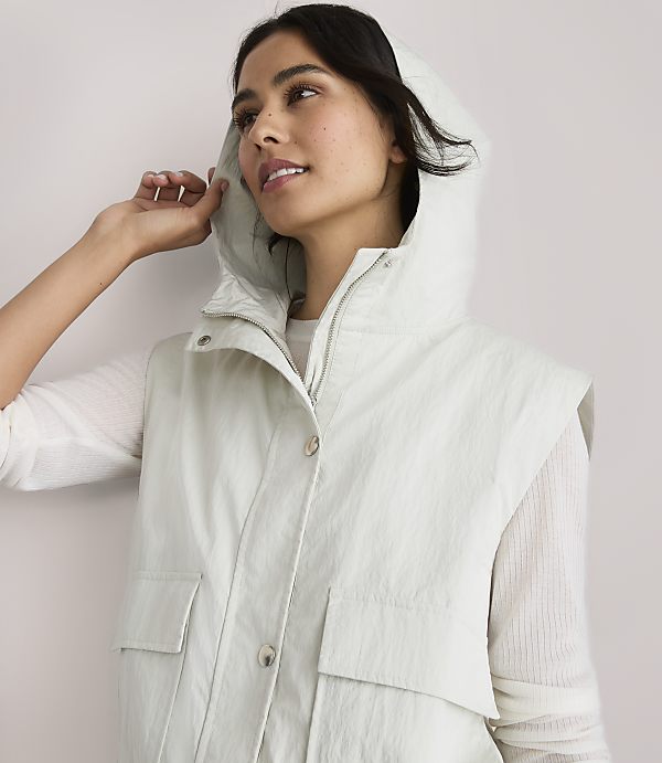 A woman in a light-colored hooded vest and green pants stands against a plain white background.