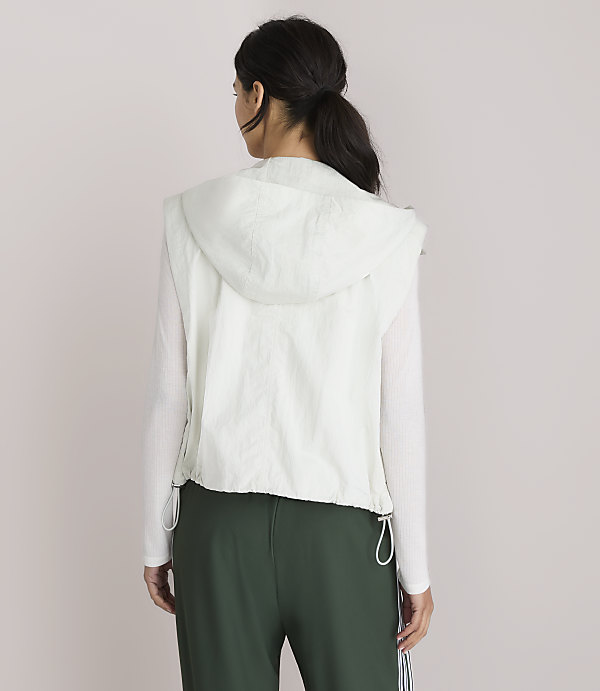 A woman in a white hooded vest and green pants stands against a plain white background.
