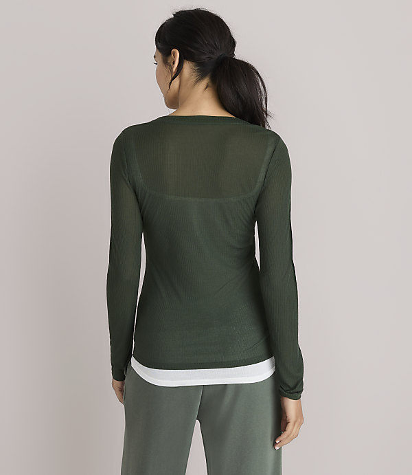 A woman in a sheer green long-sleeved shirt and white tank top stands against a plain, light gray background.
