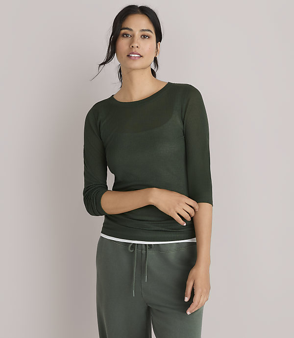 A woman in a dark green, sheer, long-sleeved shirt and green pants poses against a plain white background.