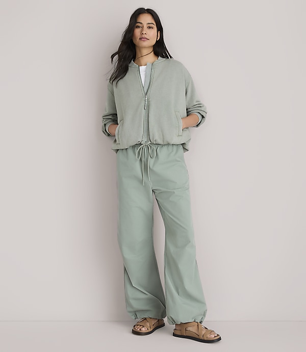 A woman stands in the foreground wearing a sage green bomber jacket and matching wide-leg pants, against a plain white background.