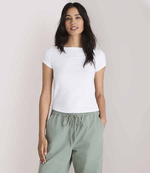 A woman in a white t-shirt and green pants stands against a plain white background.