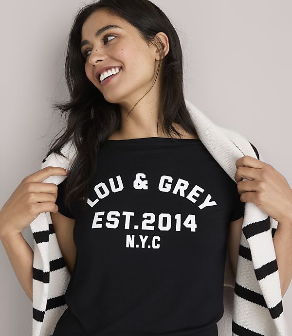 A smiling woman in a black t-shirt with "LOU & GREY EST. 2014 N.Y.C." printed on it, draped with a striped scarf, stands against a plain white background.