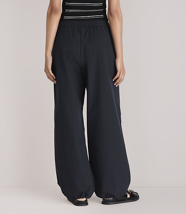 A person wearing dark, wide-leg pants and sandals stands against a plain white background.