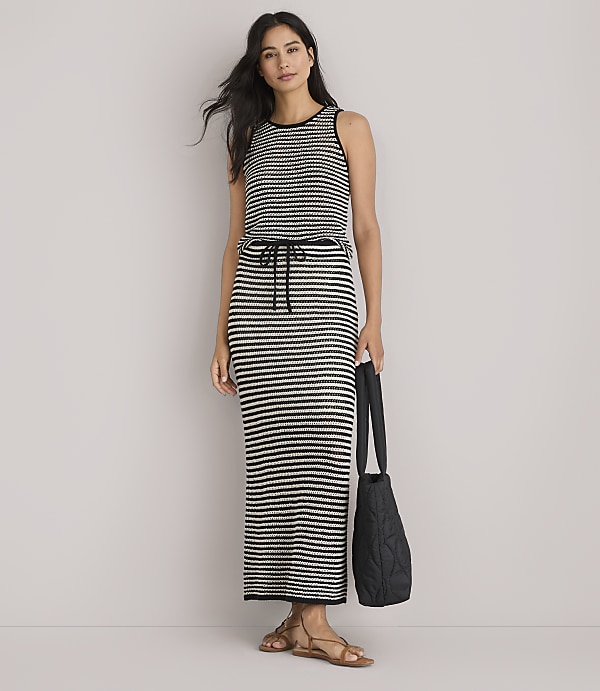 A woman in a striped maxi dress and sandals stands against a plain white background, holding a black quilted tote bag.