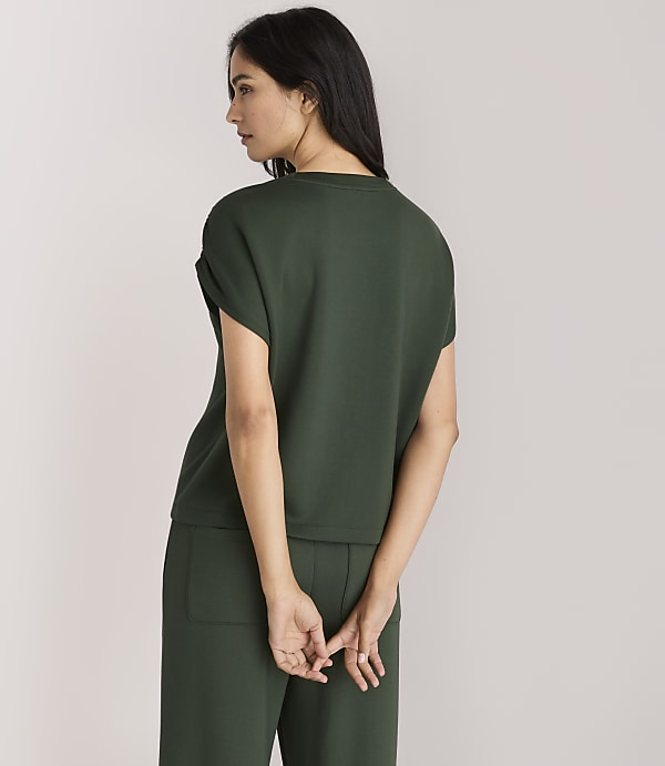 A woman in a dark green top and pants stands with her back to the viewer against a plain white background.