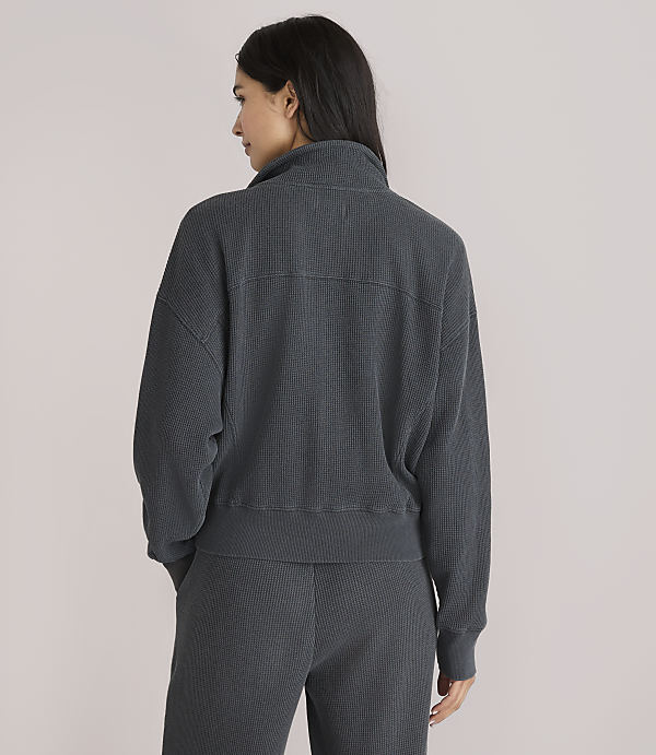 A woman in a dark grey ribbed quarter-zip sweatshirt and matching pants stands against a plain white background.