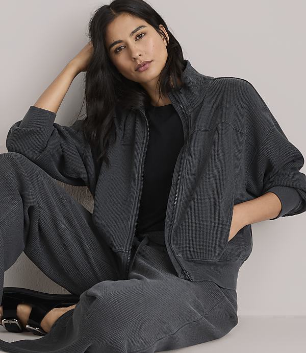 A woman in a dark grey zip-up jacket and matching pants stands against a plain white background.