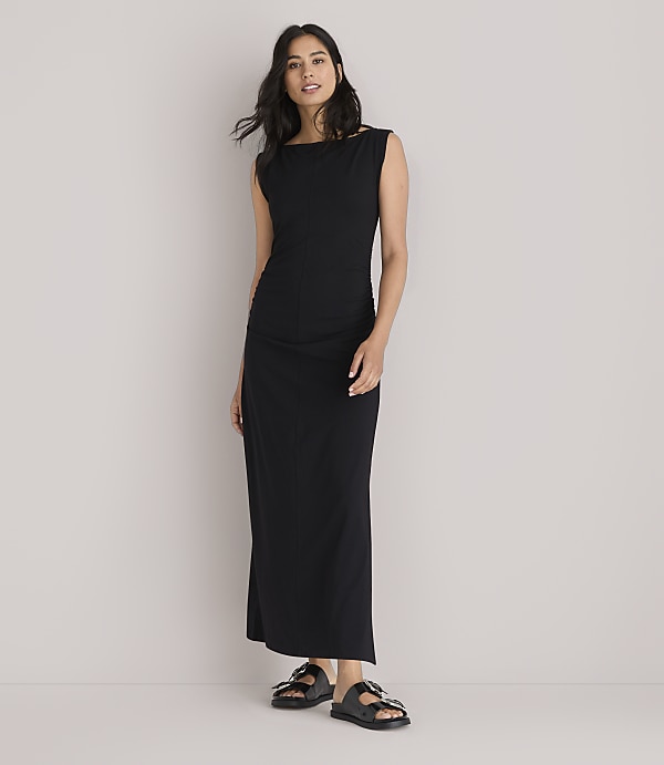 A woman in a black maxi dress and sandals stands against a plain white background.
