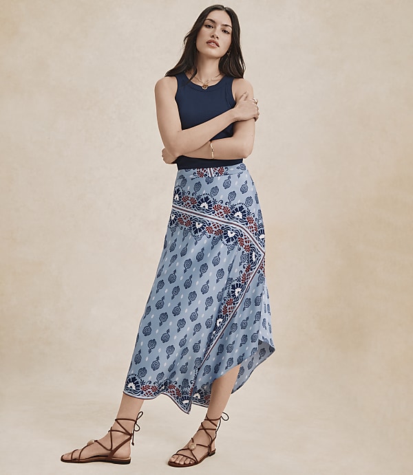 A woman in a blue tank top and a flowing, patterned skirt stands against a plain, light brown background.