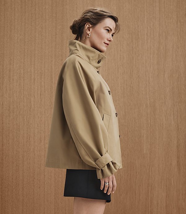 A woman in a tan jacket and black skirt stands against a warm, wood-paneled background.