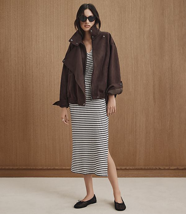 A woman in a brown jacket and striped dress stands against a wood-paneled background.