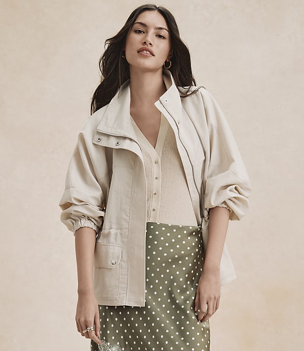 A woman in a cream jacket and green polka dot skirt stands against a textured beige background.