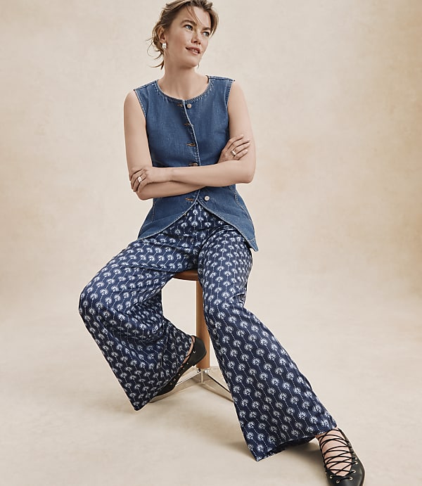 A woman in a denim vest and patterned pants sits on a stool against a plain, light-colored background.
