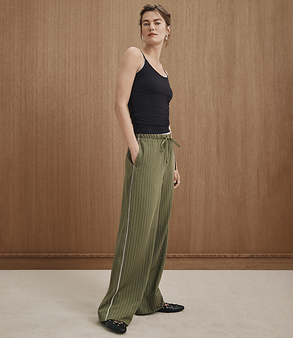 A woman in a black tank top and olive green striped pants stands against a warm, wood-paneled background.