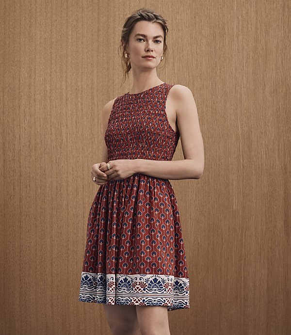 A woman in a patterned red dress stands against a textured brown background.