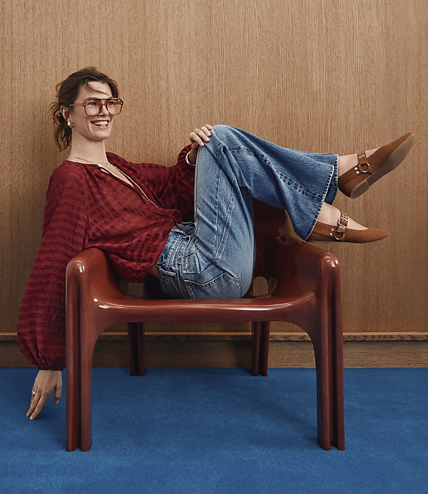 A woman in a red blouse and blue jeans sits on a brown chair against a wood-paneled wall and a blue carpeted floor.