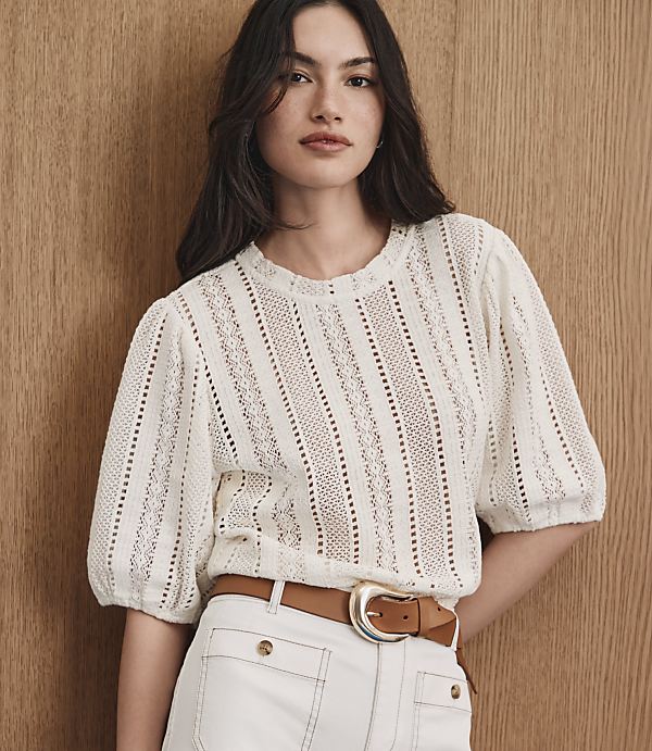 A woman in a white lace top and white pants with a brown belt stands against a wooden background.