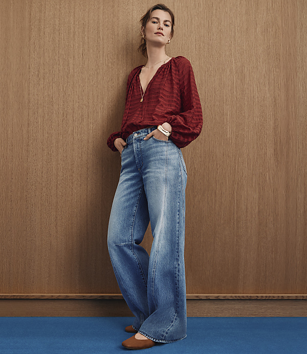 A woman in a red patterned blouse and wide-leg jeans stands against a wood-paneled wall with a blue floor in the foreground.