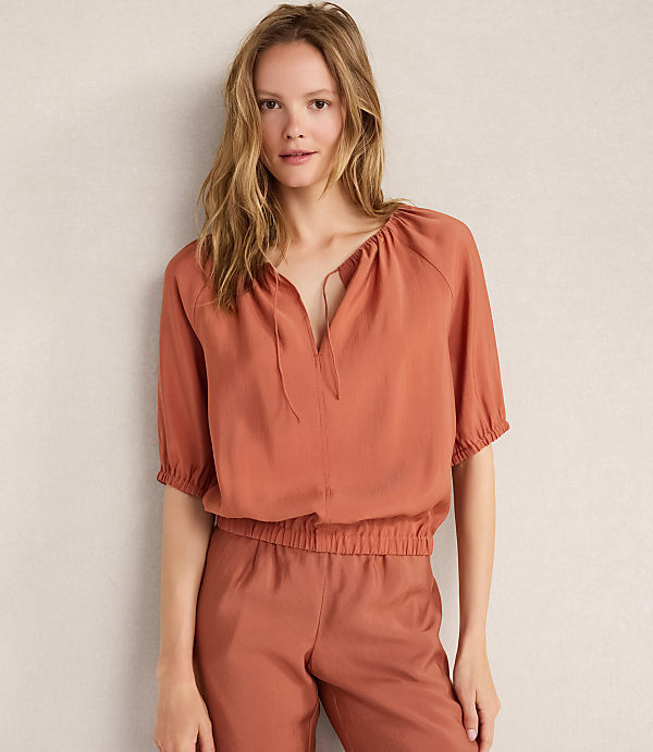 A woman in a rust-colored top and pants stands against a plain, light-colored wall.