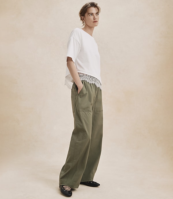 A woman in a white t-shirt and olive green pants stands against a textured, light brown background.