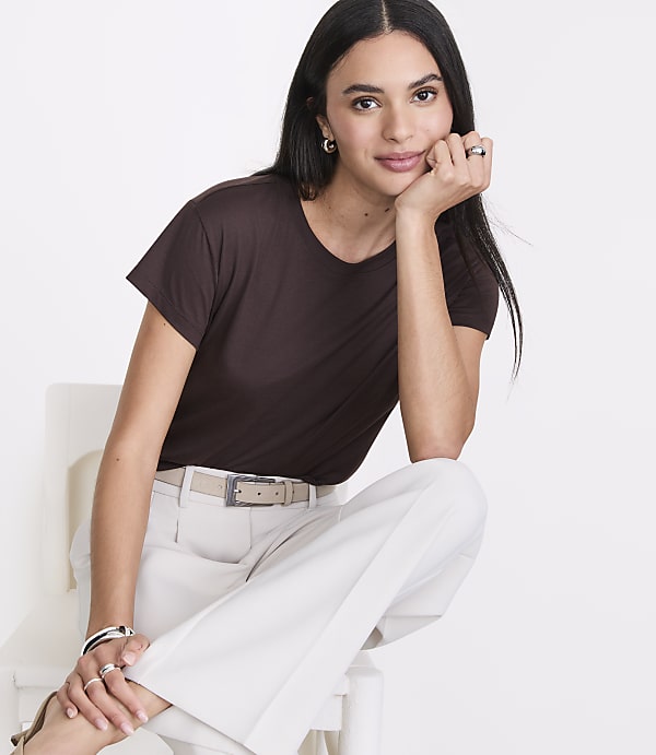 A woman in a brown t-shirt and light-colored pants rests her chin on her hand against a white background.