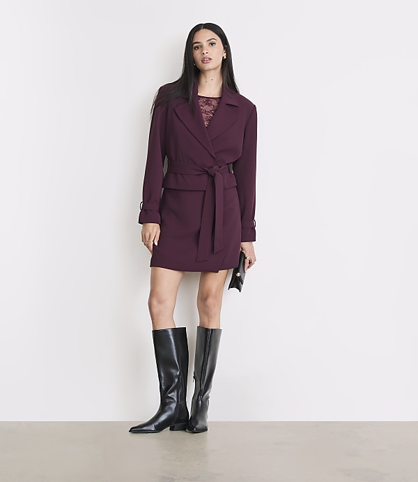 A woman in a burgundy dress and black boots stands against a plain white background.