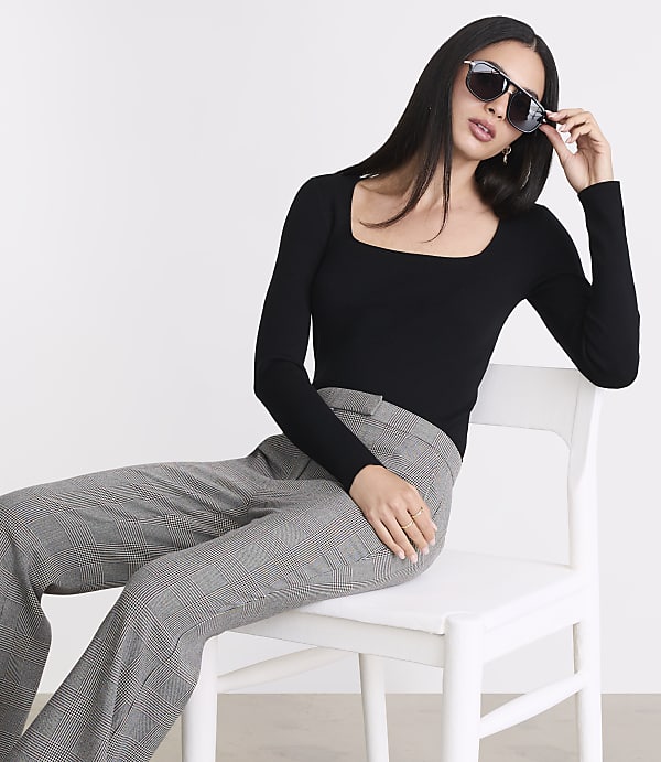 A woman in a black top and plaid pants sits on a white chair against a plain white background, adjusting her sunglasses.