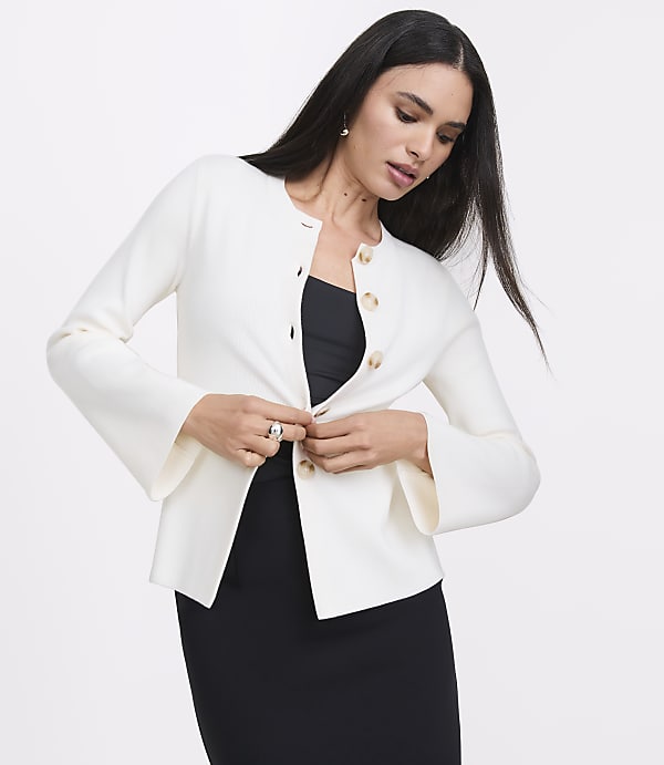 A woman in a white cardigan with bell sleeves buttons it over a black dress against a plain white background.