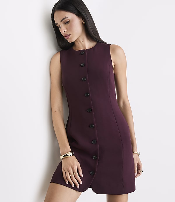 A woman in a plum-colored sleeveless dress with a button-down front stands against a plain white wall.