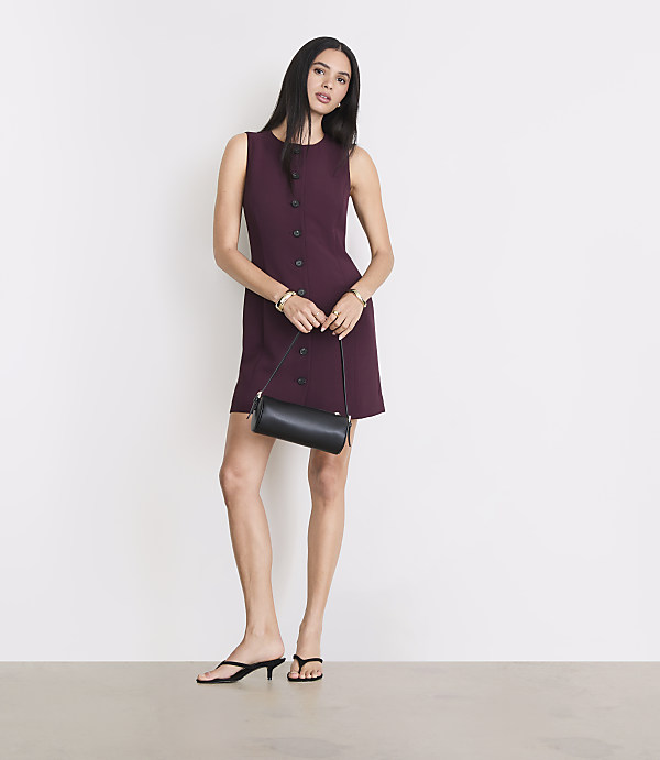 A woman in a burgundy dress and black heels stands with a black purse against a plain white background.