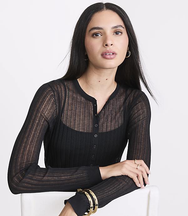 A woman with long dark hair and hoop earrings, wearing a sheer black cardigan over a black top, leans forward against a white surface with a plain white background.