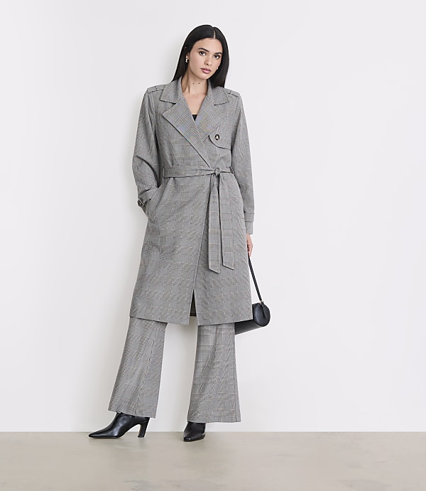 A woman in a plaid trench coat and matching pants stands against a plain white background, holding a black purse.