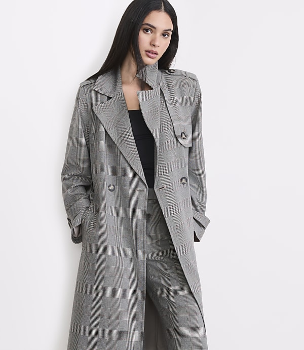 A woman in a plaid trench coat and pants stands against a plain white background.