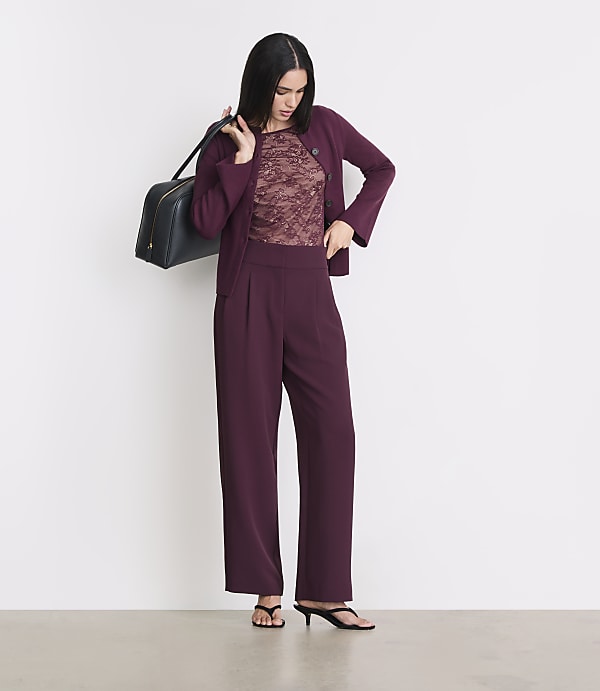 A woman in a burgundy suit and lace top stands with a dark bag over her shoulder against a plain white background.