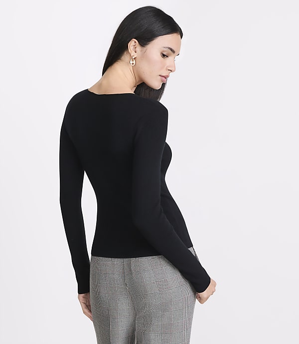 A woman in a black long-sleeved shirt and plaid pants stands against a plain white background.